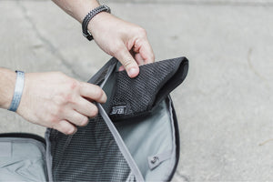 Person placing a black textured pouch into a gray bag on a neutral background