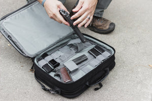 Person handling a gun in an open black suitcase on a concrete surface