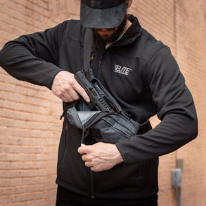 Person in black jacket with 'ELITE' logo holding a tactical bag against a brick wall.