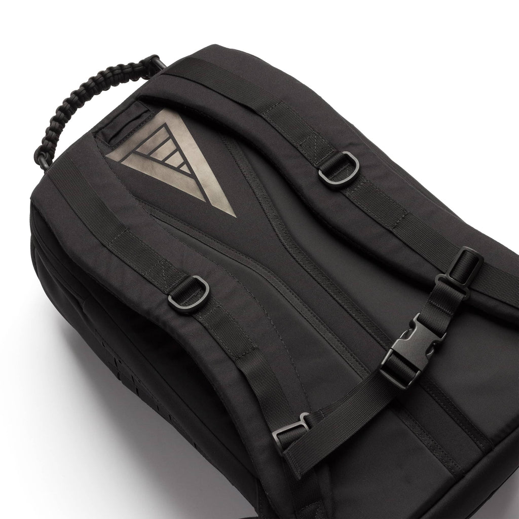A black nylon backpack with a triangular metallic silver logo patch on the front, black straps, and plastic buckles, shown on a clean white background.