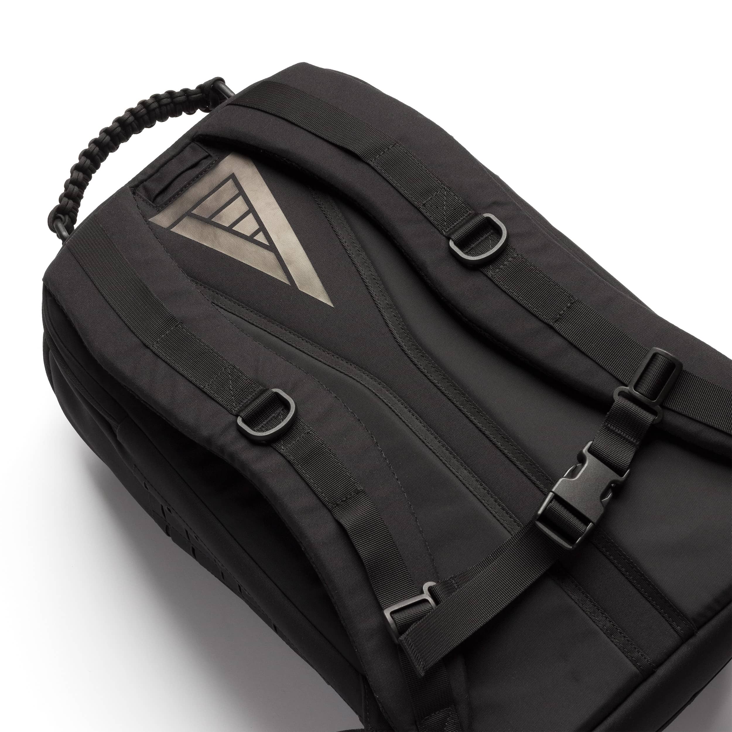 A black nylon backpack with a triangular metallic silver logo patch on the front, black straps, and plastic buckles, shown on a clean white background.
