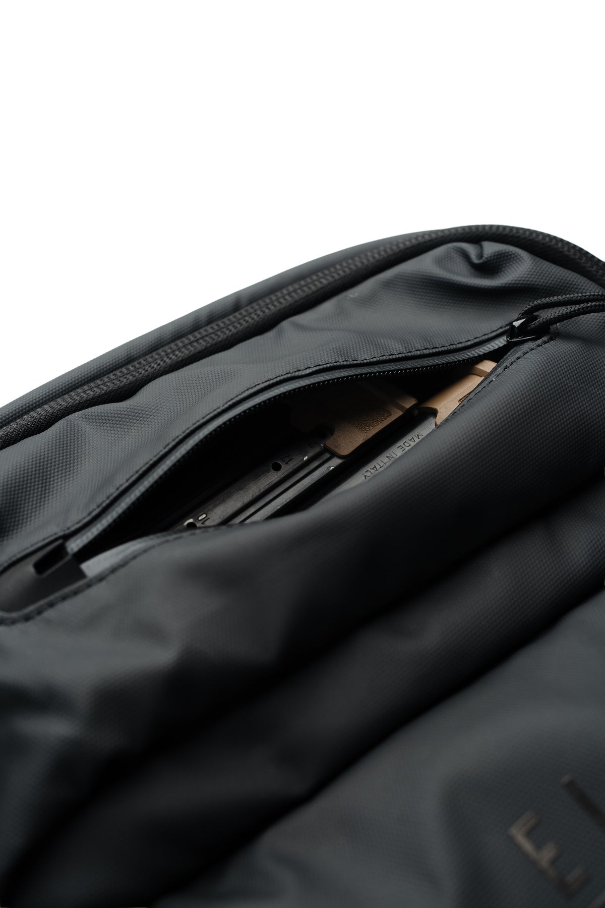 Close-up of a black nylon bag with a partially unzipped pocket, showing a brown paper sleeve or document inside, on a white background.