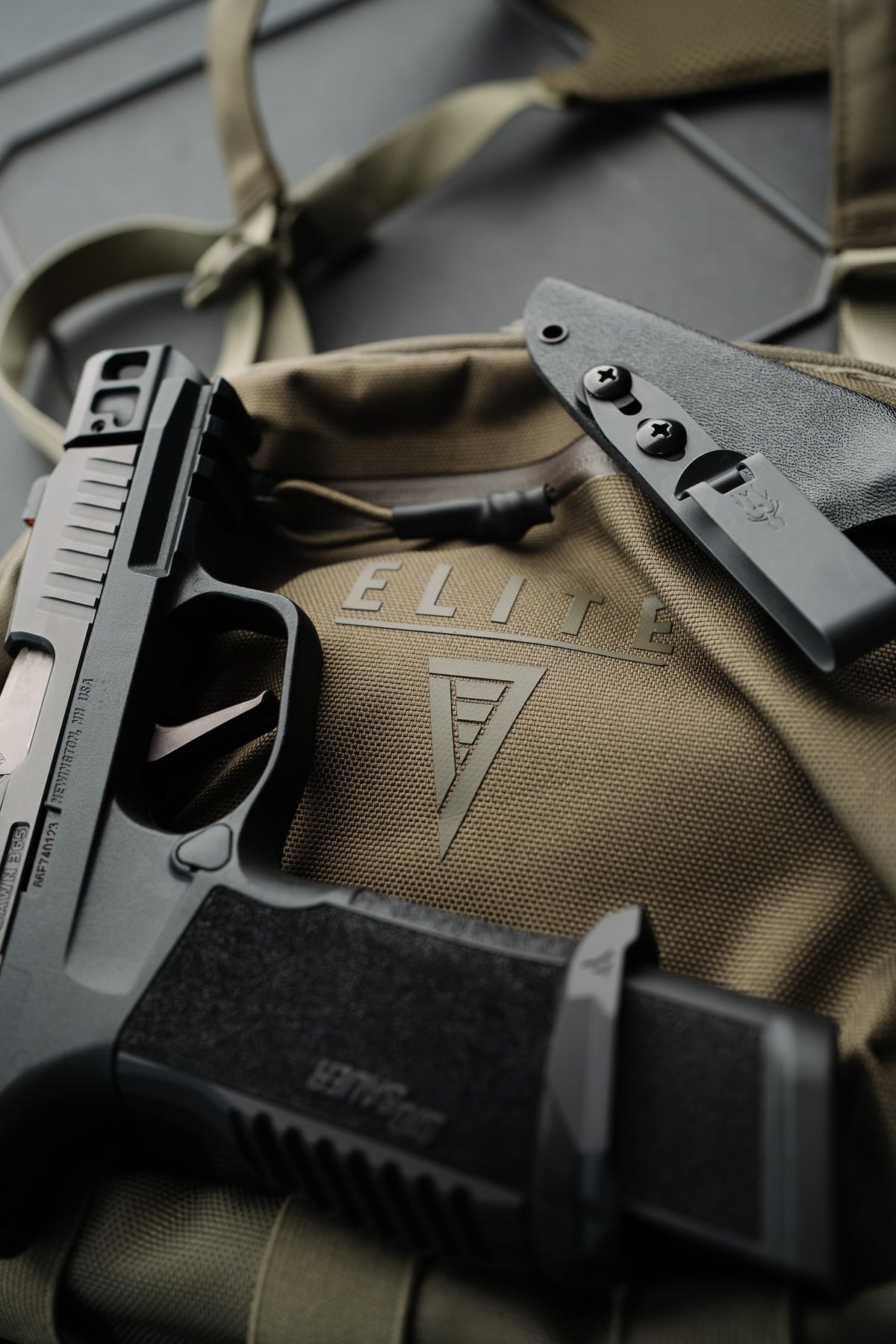 A black semi-automatic pistol and a black sheath are arranged on top of a tan tactical backpack with the brand name ELITE and a triangular logo embossed on the fabric.