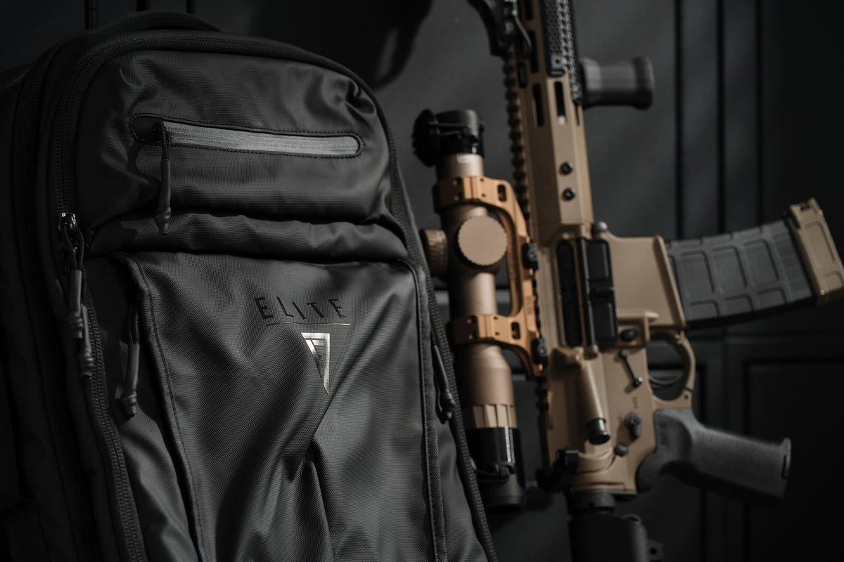 A black tactical backpack with the ELITE brand logo on the front pocket, shown next to a tan and black rifle with a mounted scope in a dark setting.