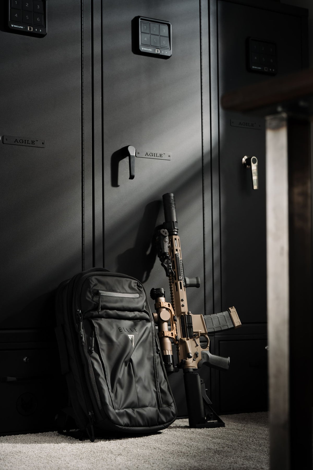 A black tactical backpack stands on a carpeted floor next to a tan and black rifle, positioned in front of dark metal storage lockers.