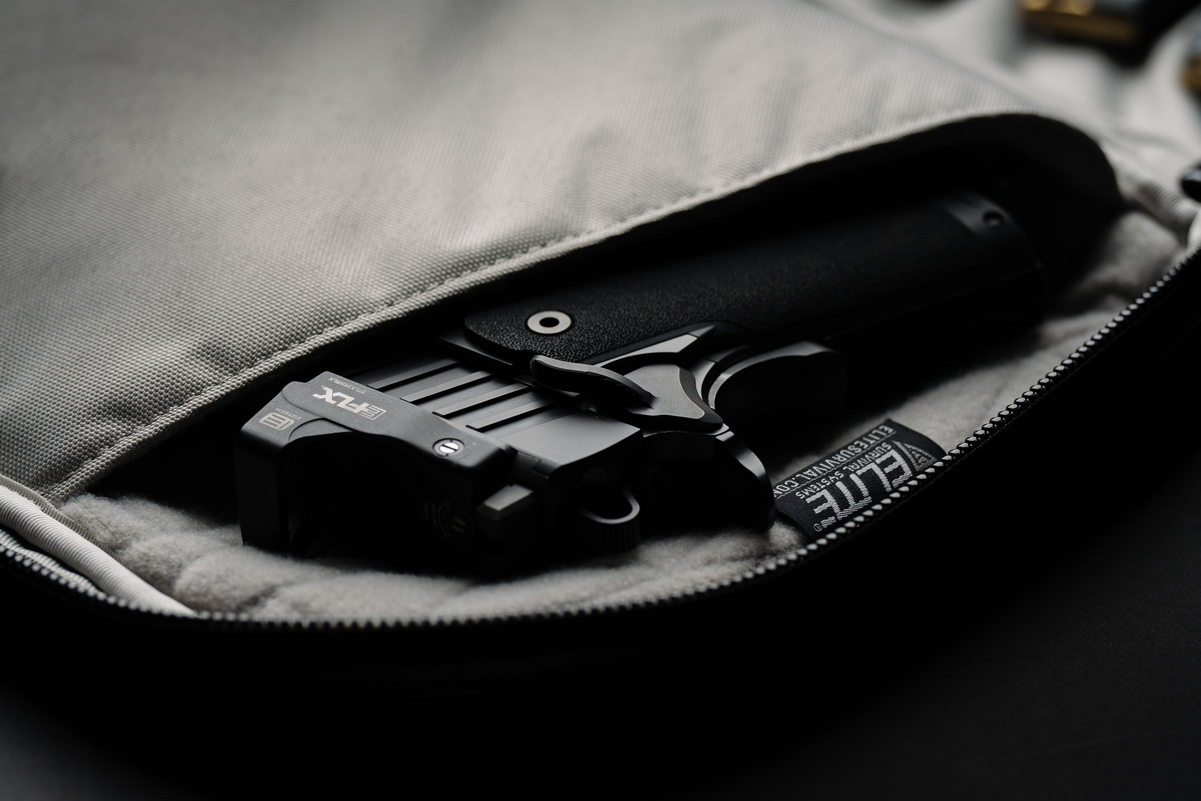 A black tactical handgun with a black holster and a black vertical magazine insert, shown partially tucked into an open gray zippered bag. A black fabric tag on the holster reads 'ELITE'.