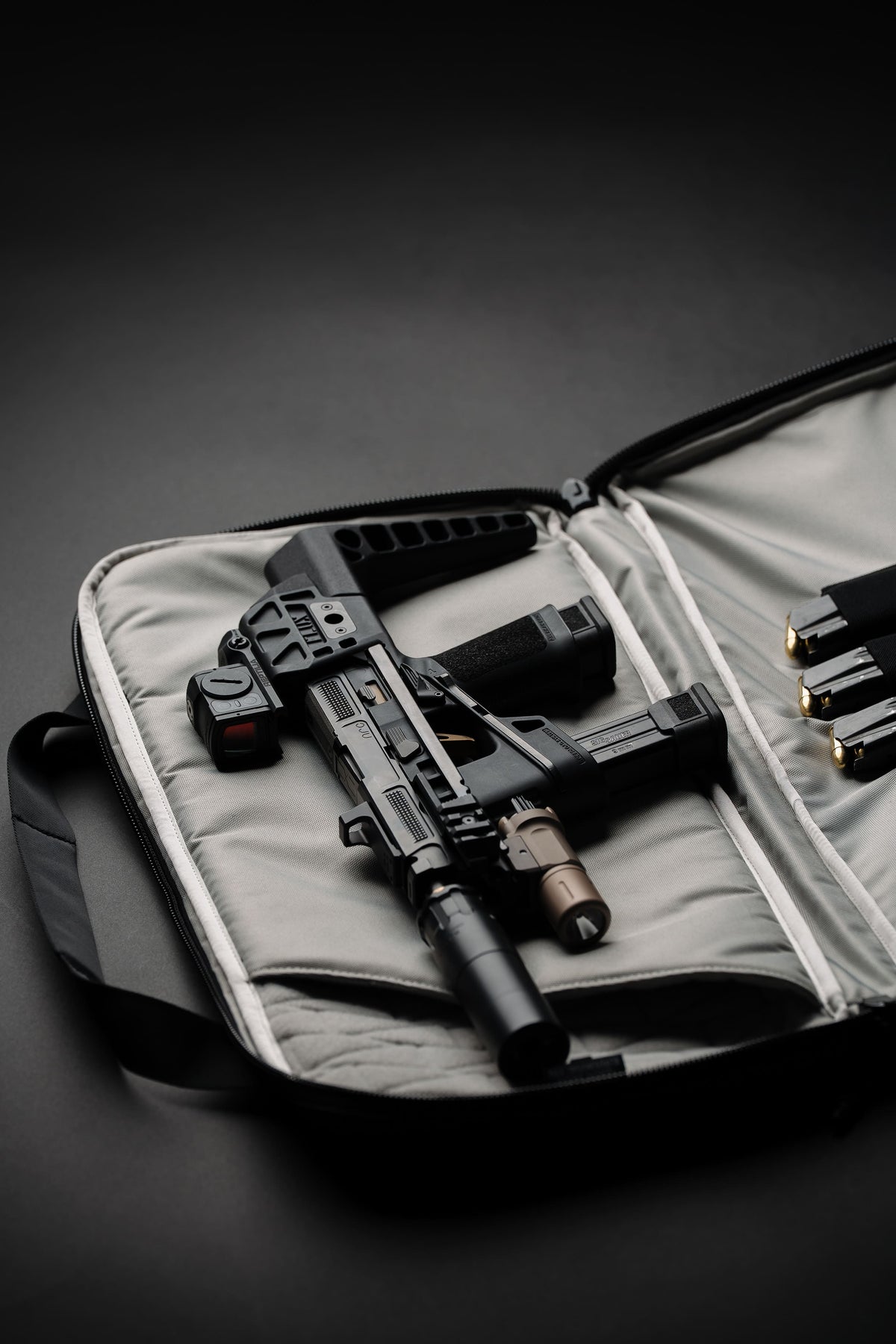 A black tactical rifle with an underbarrel attachment is displayed inside an open padded gun case on a dark studio background. Several magazines are visible in the case, and the scene is lit with dramatic, moody lighting.