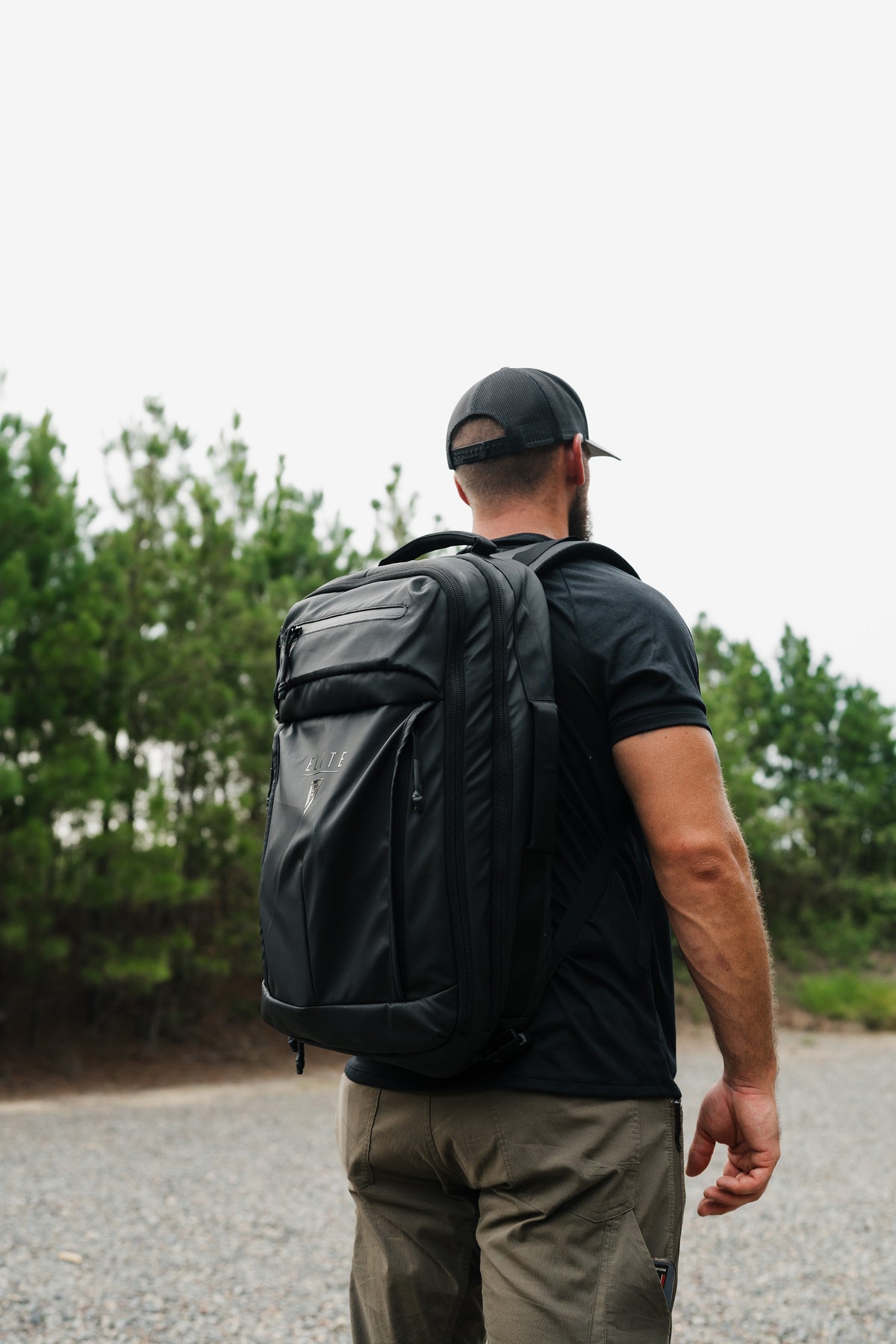 A man viewed from behind wearing a black backpack, black t-shirt, and khaki pants while walking outdoors on a gravel path with trees in the background.