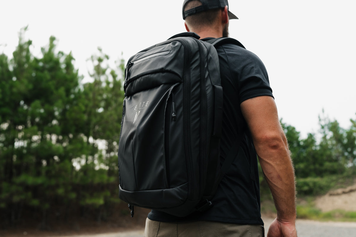 A man wearing a black travel backpack with multiple zippered compartments, shown from behind in an outdoor setting with trees in the background.
