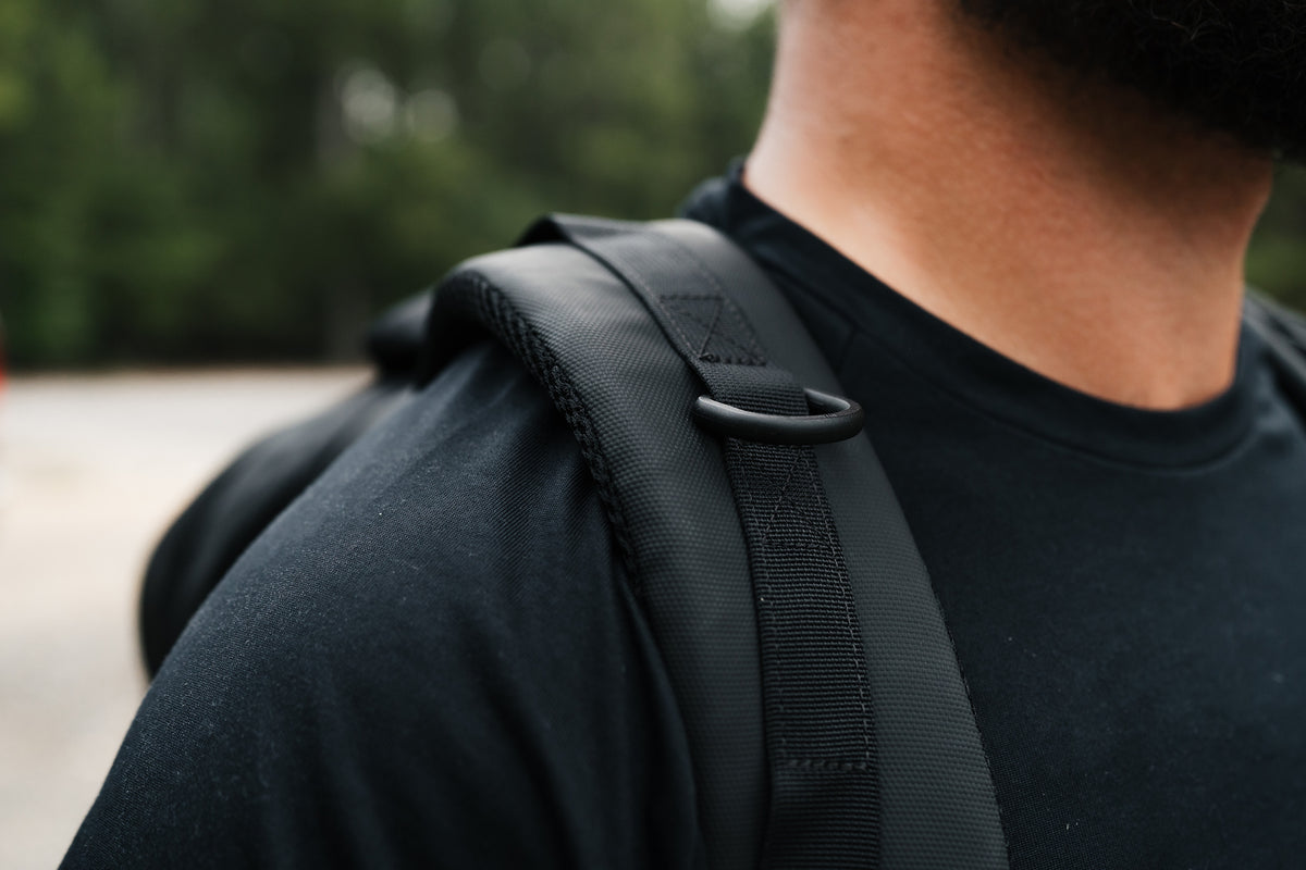 Close-up of a person wearing a black backpack, showing the shoulder strap and black D-ring hardware over a black t-shirt, with a blurred outdoor background.