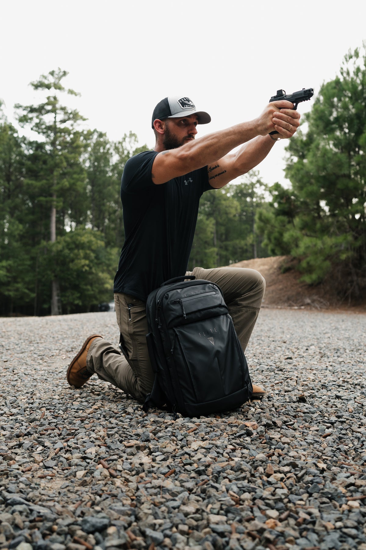 A man kneels on a gravel surface outdoors, holding a handgun with both hands. A black backpack sits beside him, and trees are visible in the background.