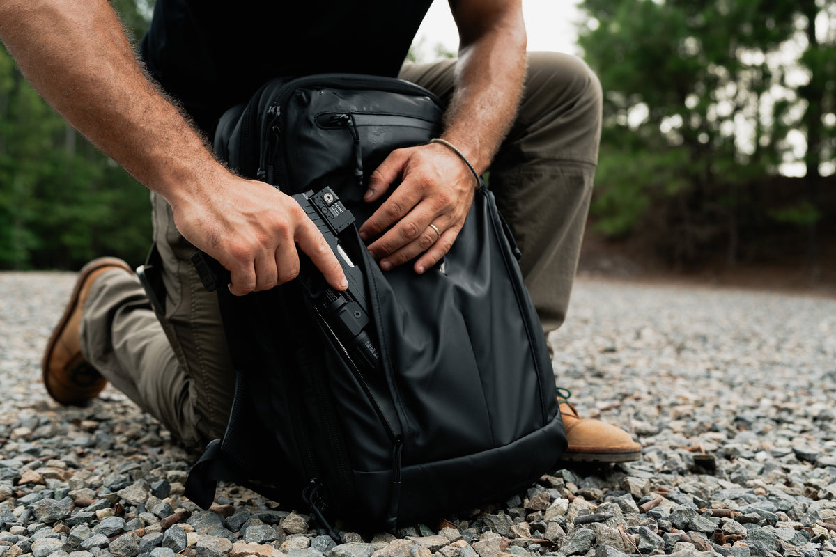 A black backpack with a side pocket containing a handgun, shown on a gravel surface outdoors with a person kneeling beside it.
