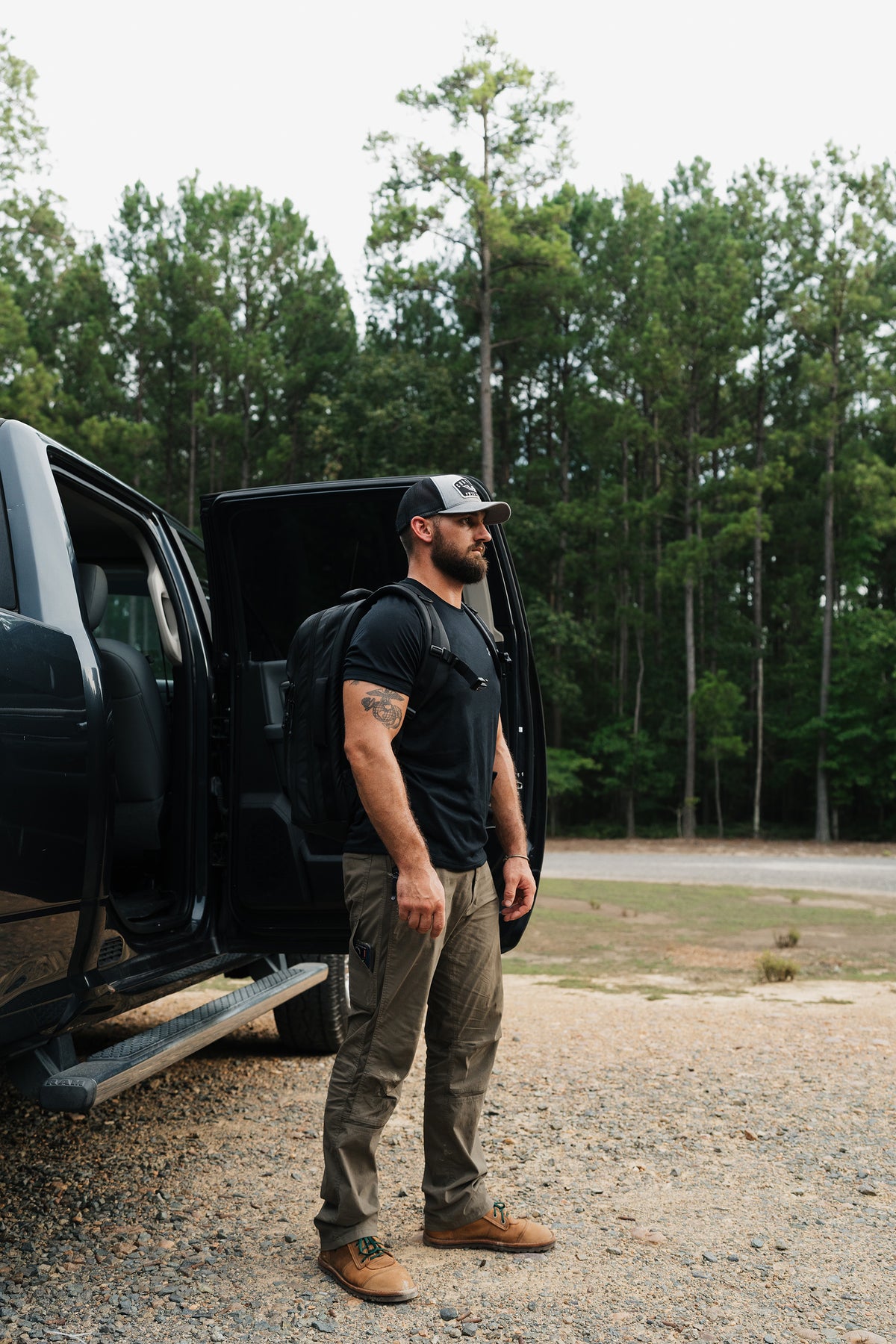 A man wearing a black t-shirt, olive cargo pants, and a black baseball cap stands next to an open black truck door in an outdoor gravel setting with a forest background.
