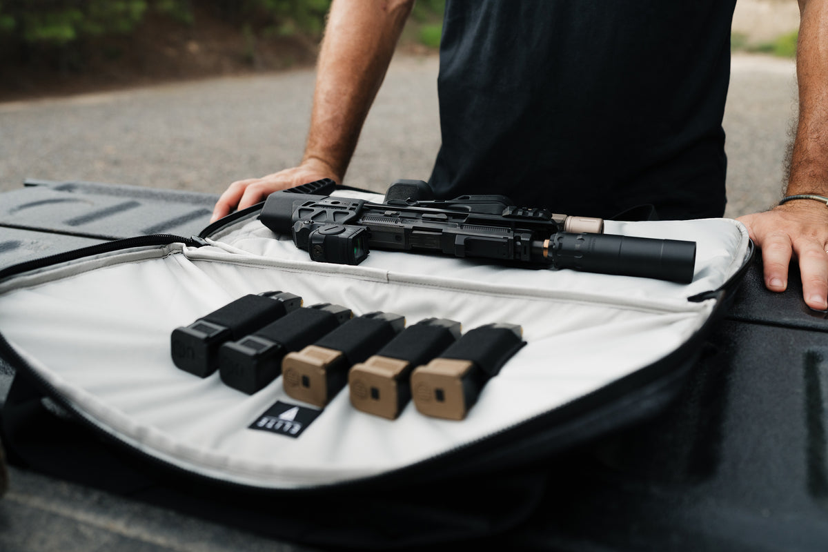 A black tactical rifle with a mounted flashlight and suppressor rests on an open white and black gun case, alongside several magazine inserts. A person in a black shirt is visible from the torso down, standing outdoors on a truck bed.