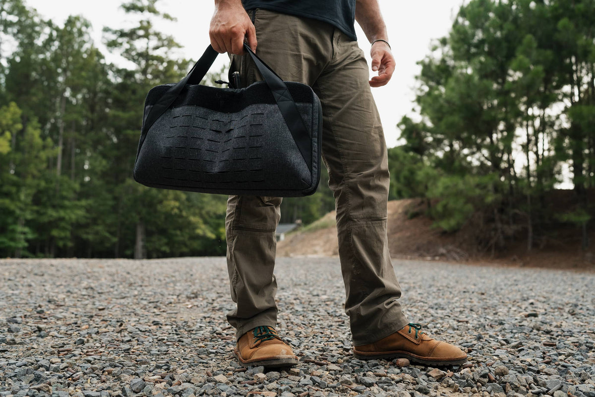 A person outdoors on a gravel path wearing olive cargo pants and brown shoes, holding a black textured duffel bag.