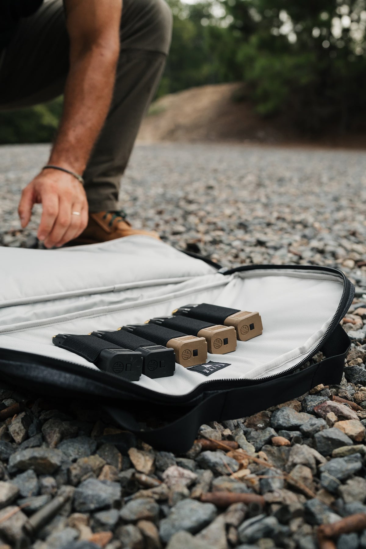 A black zippered carrying case opened on a gravel path outdoors, holding several black and tan rectangular accessories. A person's hand and lower leg are visible kneeling beside the case, with trees and a dirt mound blurred in the background.