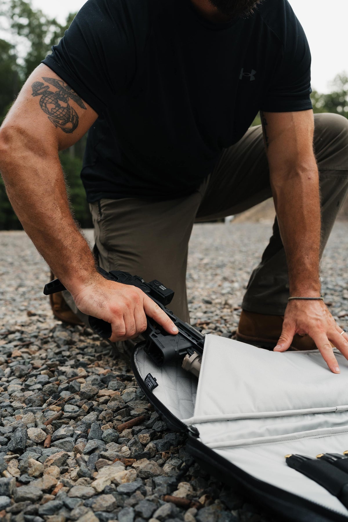 A man kneels on a gravel surface, placing a black handgun into an open black and silver gun case. He is wearing a black Under Armour t-shirt and khaki pants.