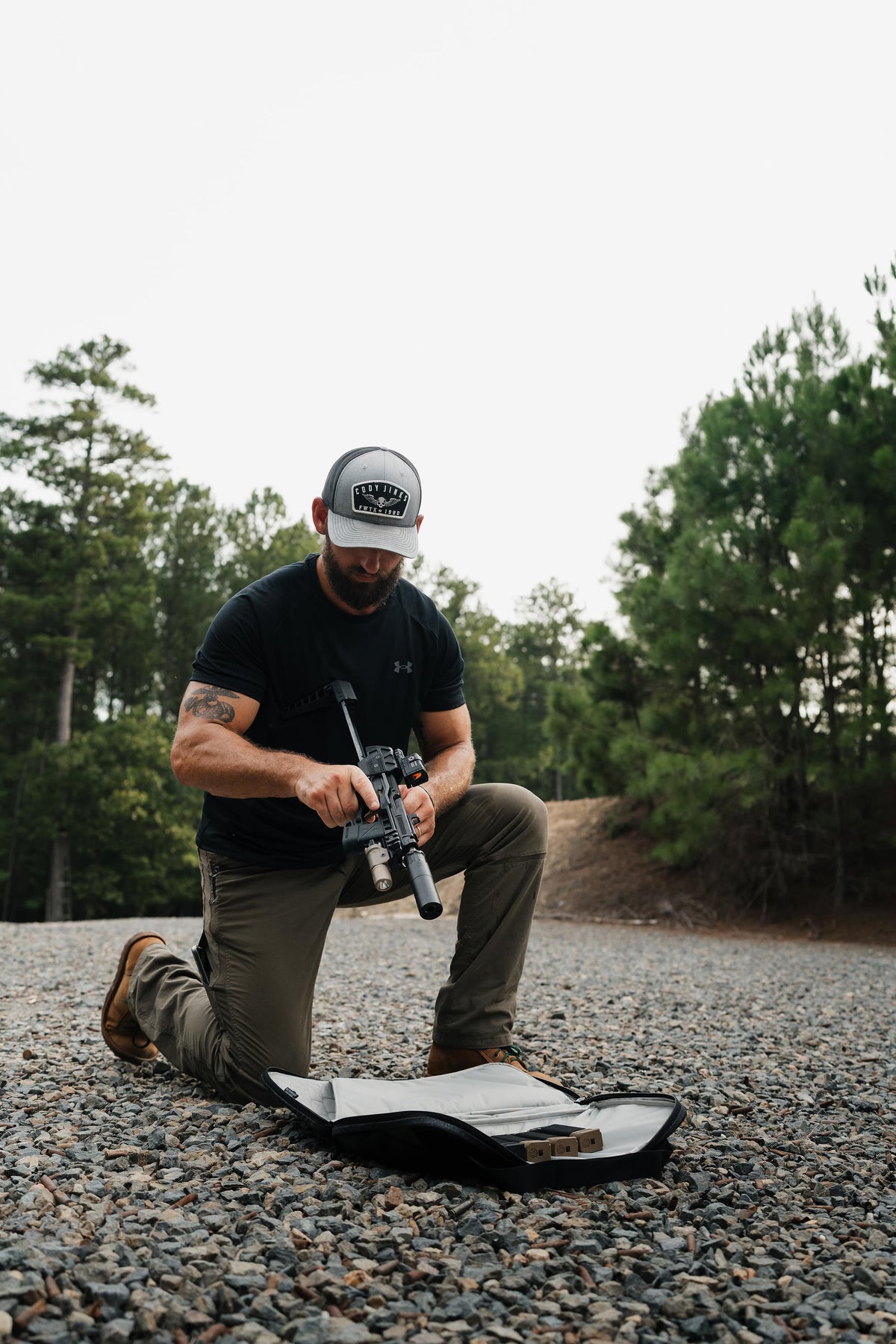 A man kneels on a gravel surface outdoors, handling a rifle with a scope attached while an open black case lies in front of him. Trees and an overcast sky are visible in the background.