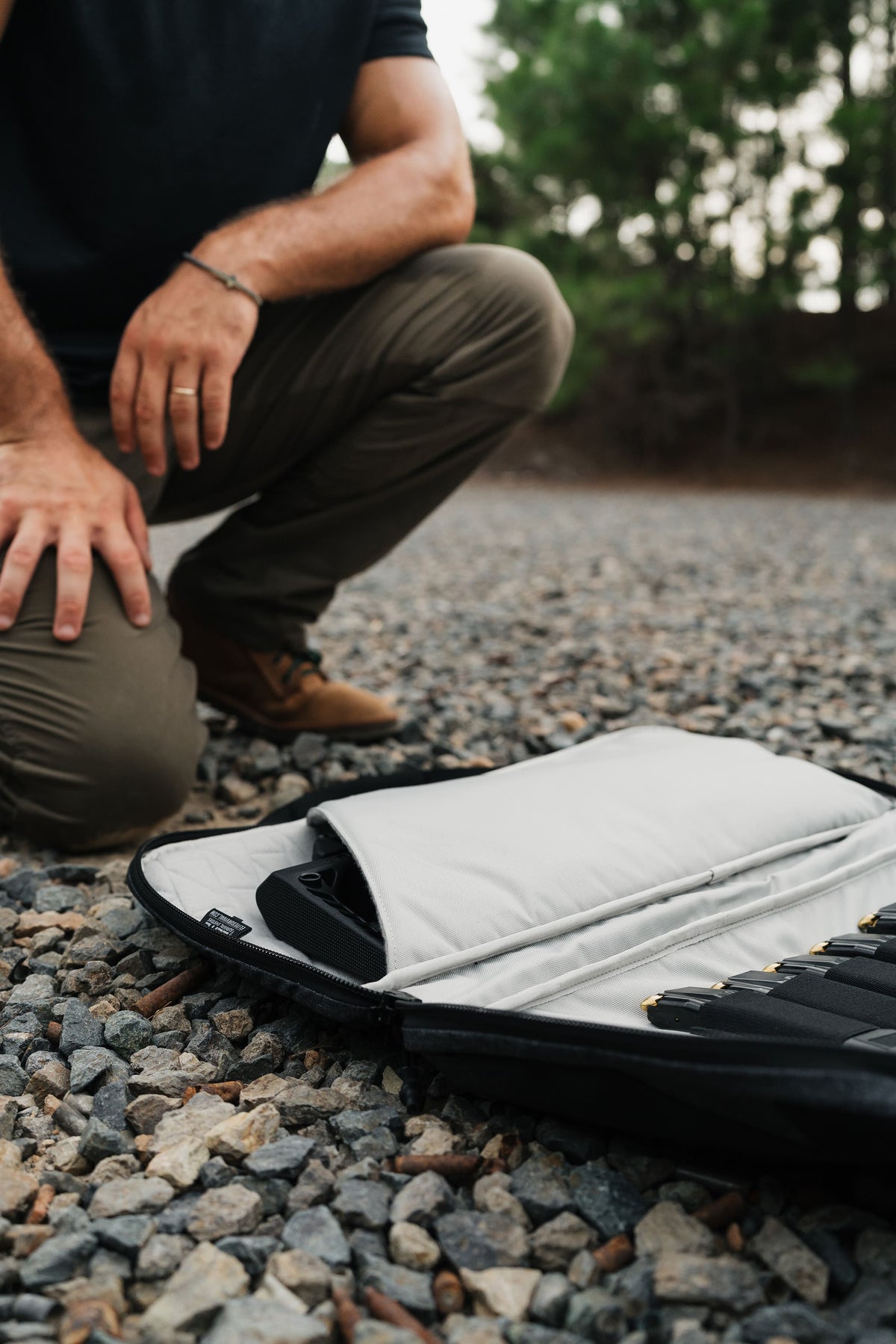 A close-up outdoor shot of a person crouching beside an open black and white padded case on a gravel surface, with trees blurred in the background.
