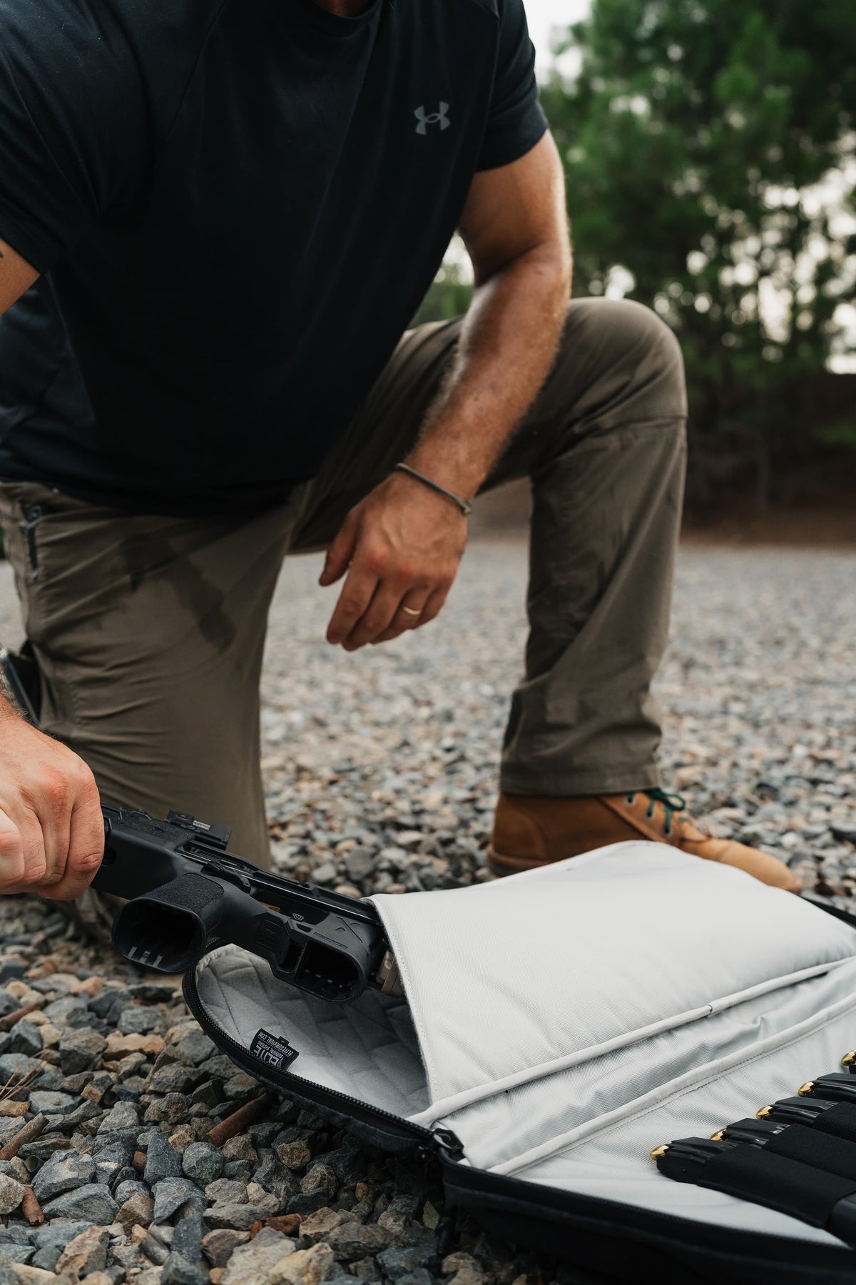 A man kneels on gravel outdoors, placing a black handgun into an open black and silver gun case with a light gray interior. The man wears a black Under Armour t-shirt and khaki pants.