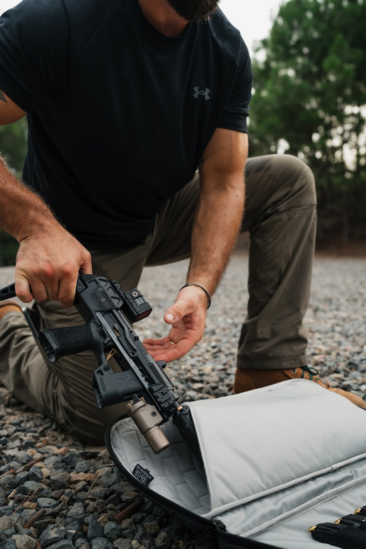A man kneels on a gravel surface outdoors, holding a black rifle above an open gray soft case. He is wearing a black Under Armour t-shirt and tan pants, with trees blurred in the background.