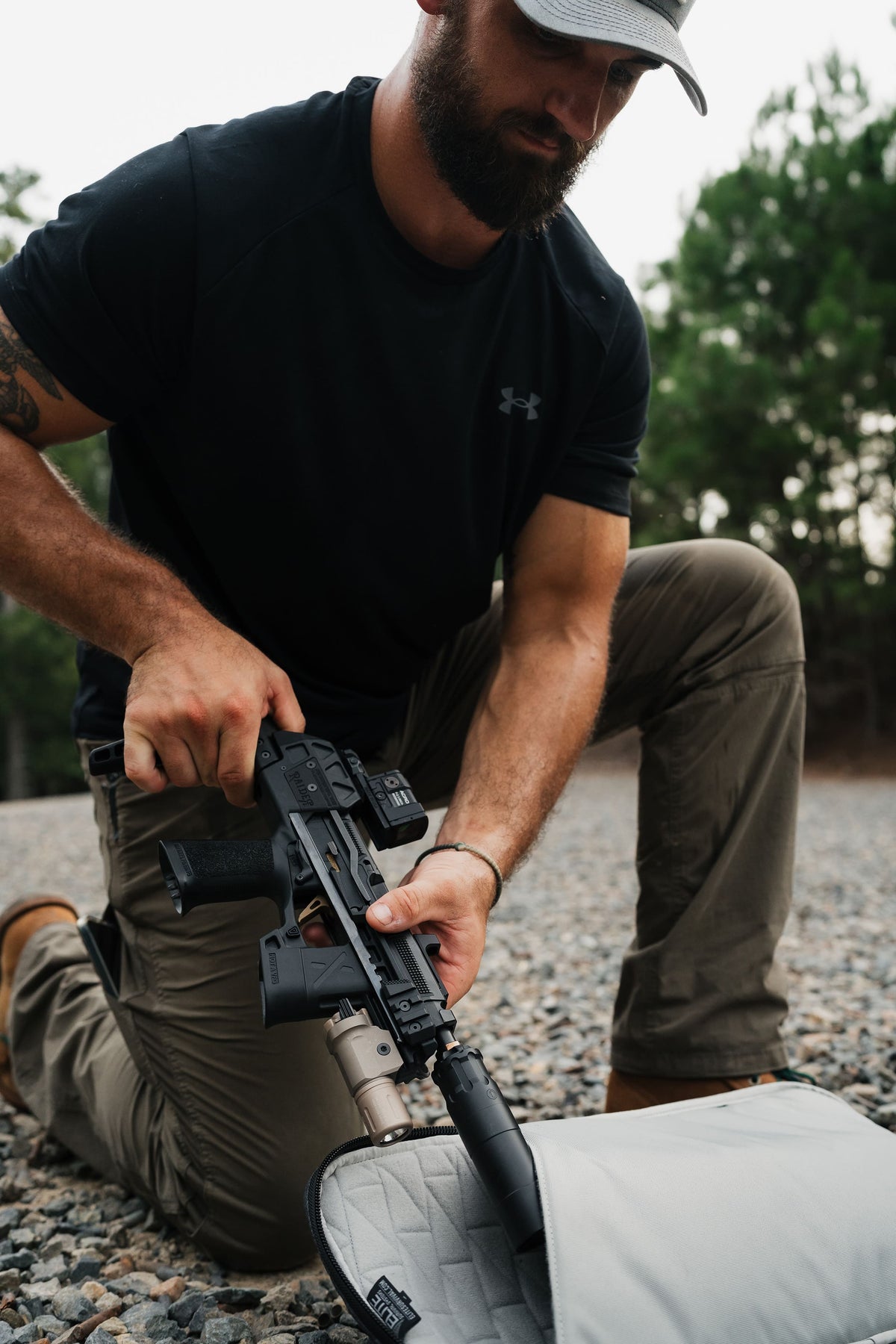 A bearded man in a black Under Armour t-shirt and khaki pants kneels on a gravel surface outdoors, holding a black tactical rifle with a mounted optic. A light gray padded case is open in the foreground.