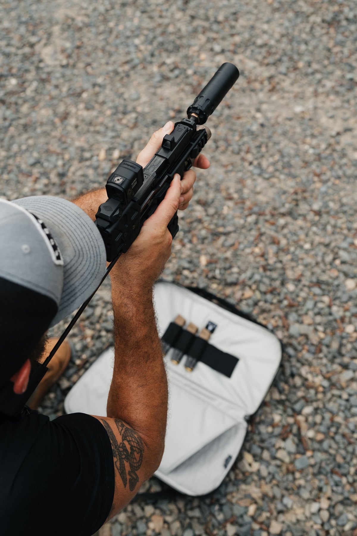 A person holding a black tactical-style firearm with a mounted optic and suppressor, photographed from above on gravel with a white case nearby.