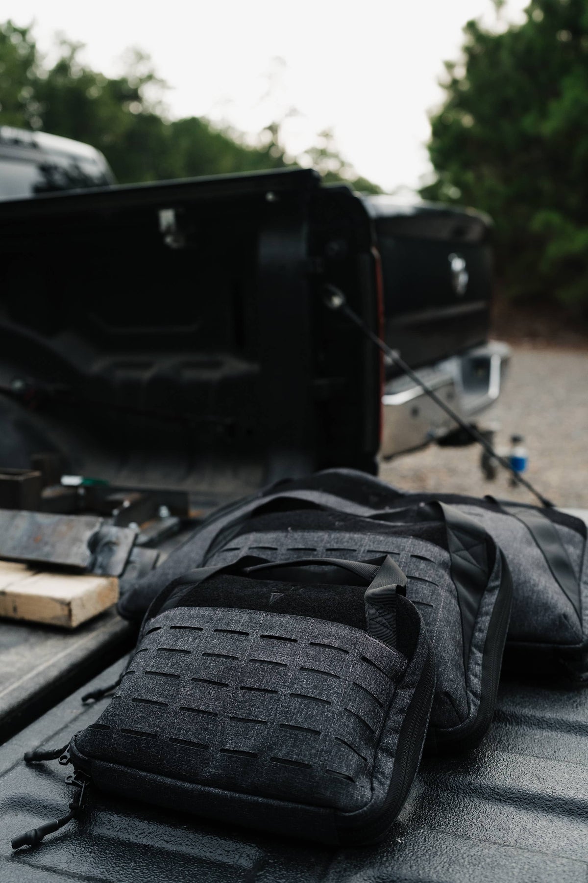 Black tactical gear bags with a textured webbed front panel resting on the tailgate of an open black pickup truck, with trees and an outdoor setting blurred in the background.