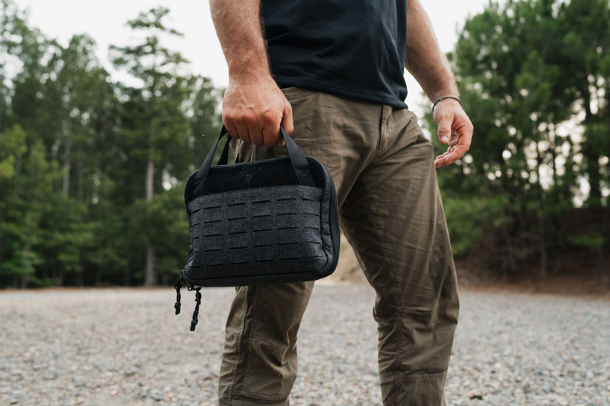 A man outdoors holding a black textured tactical pouch with MOLLE webbing and a top handle.