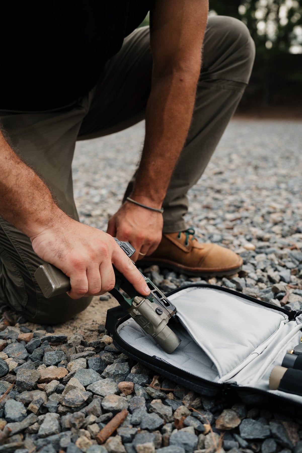A person crouching outdoors on gravel, holding a compact firearm accessory near an open black and gray case.