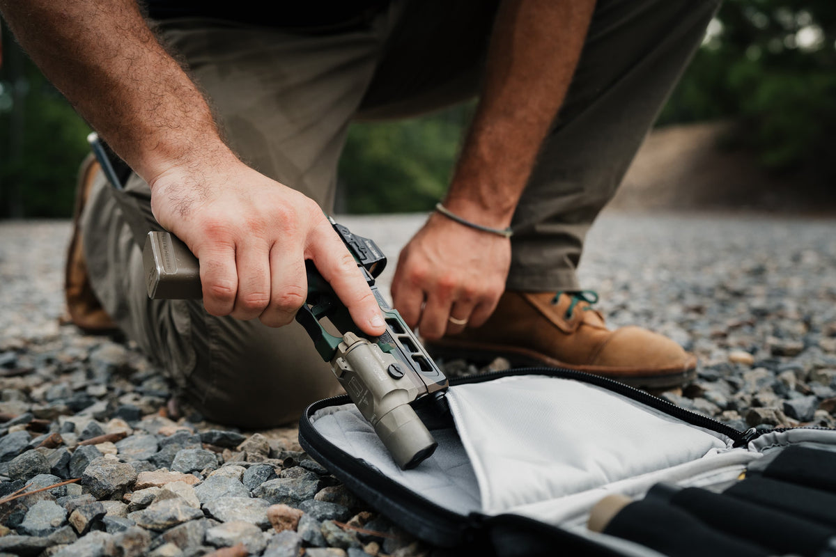 A close-up lifestyle shot of a person kneeling on a gravel surface, holding a tactical flashlight with a mounted optic over an open black and gray carrying case.
