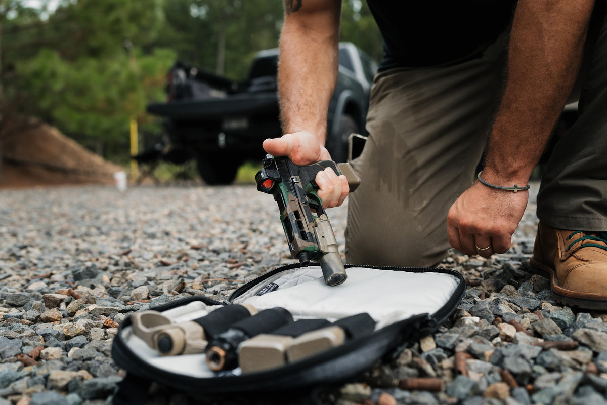 A person kneels on a gravel surface outdoors, holding a handgun above an open black case containing multiple tan and black firearm accessories. A blurred truck and trees are in the background.