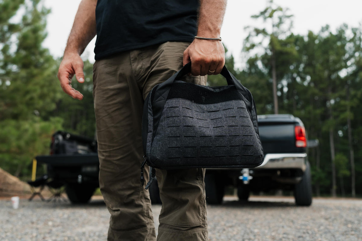 A person outdoors holding a dark gray and black textured shoulder bag with a MOLLE webbing pattern, with a truck and trees in the blurred background.