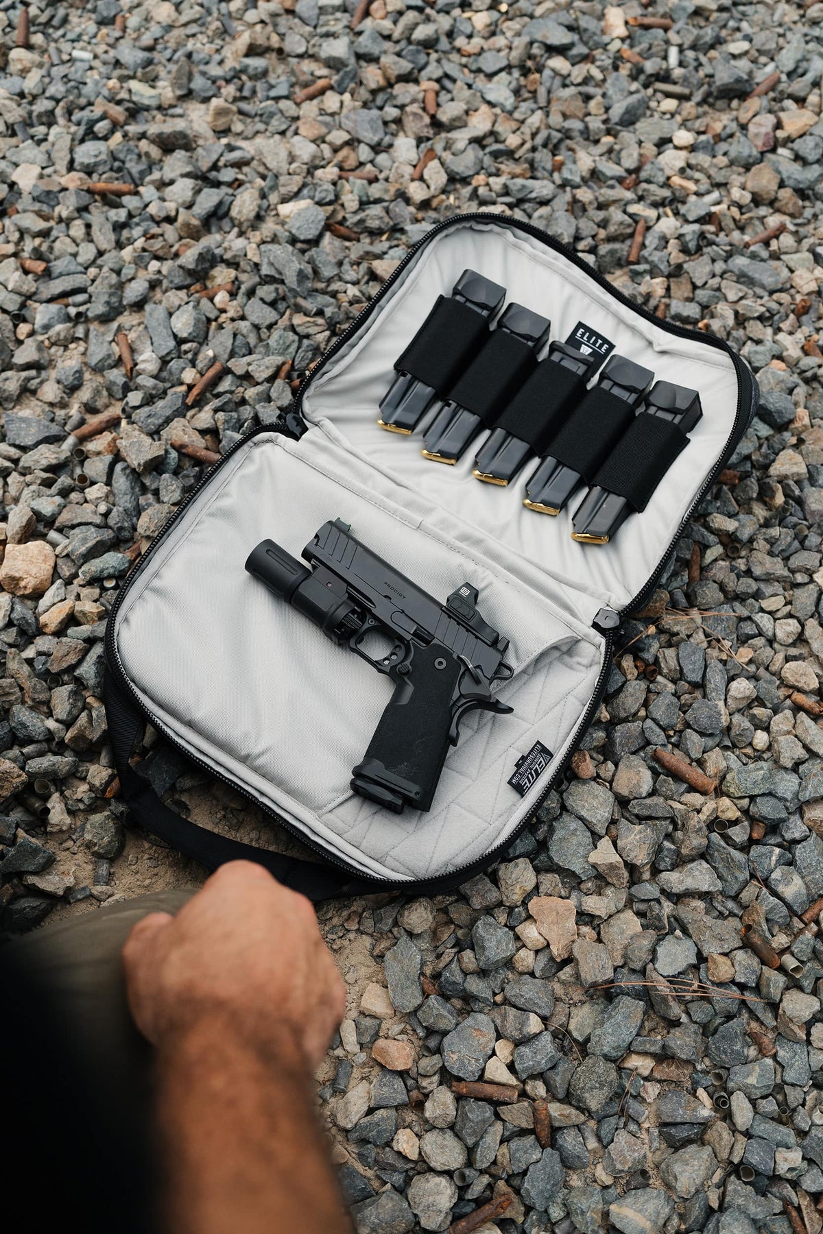 An open black and gray soft case laid on gravel, holding a black handgun and four magazine inserts. A person's hand is visible at the bottom left edge.