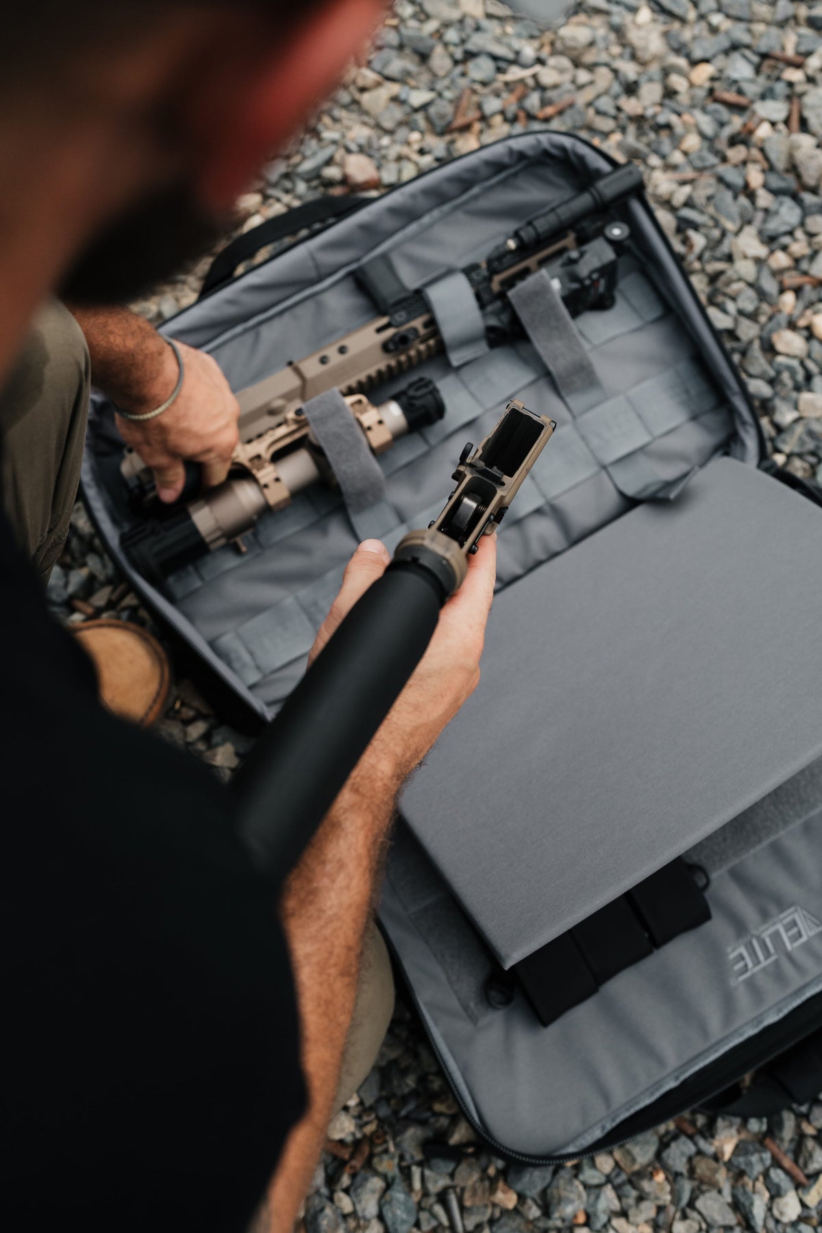 A close-up, high-angle view of a person outdoors on gravel, holding a black foam training prop in front of an open gray carrying case containing rifles. The case has the brand name ELITE visible on the lower right flap.
