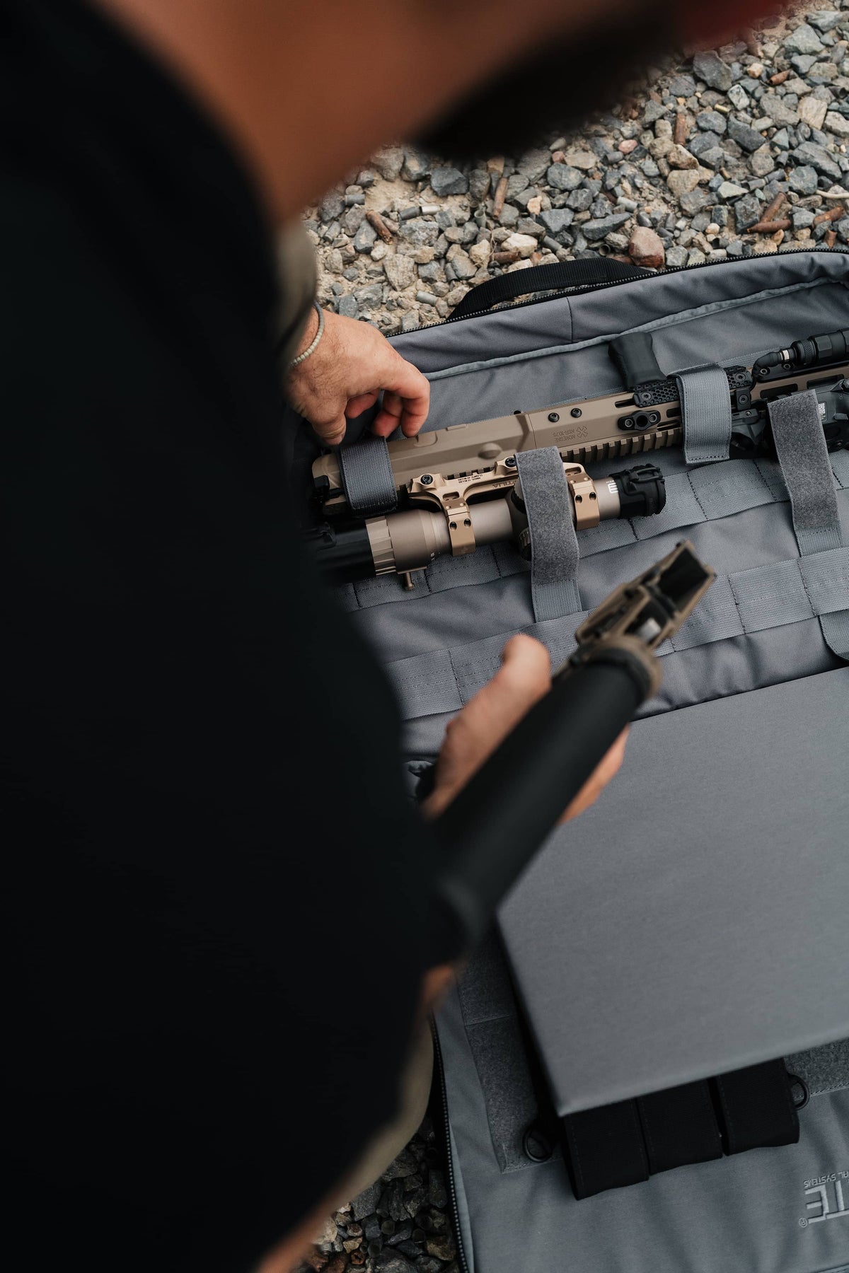 A close-up action shot of a person organizing tactical gear in a gray soft-sided case on a gravel surface. The person is holding a black cylindrical item and reaching toward a tan and black rifle mounted in the case.