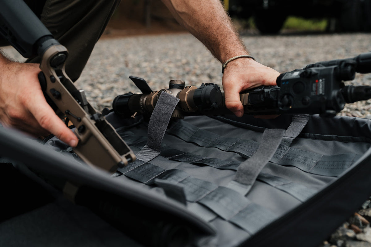 A close-up outdoor shot of a person mounting a rifle scope onto a black tactical gear bag with gray webbing straps, set on a gravel surface.