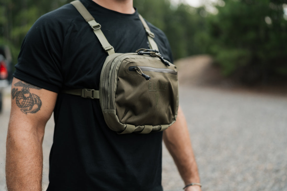 A man wearing a black t-shirt and standing outdoors on a gravel path wearing an olive green tactical chest bag.