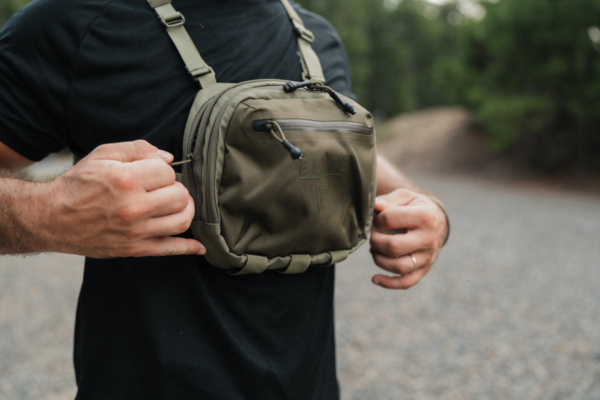 A man wearing a black t-shirt and adjusting an olive green tactical crossbody bag with a front zip pocket and a subtle embossed logo.