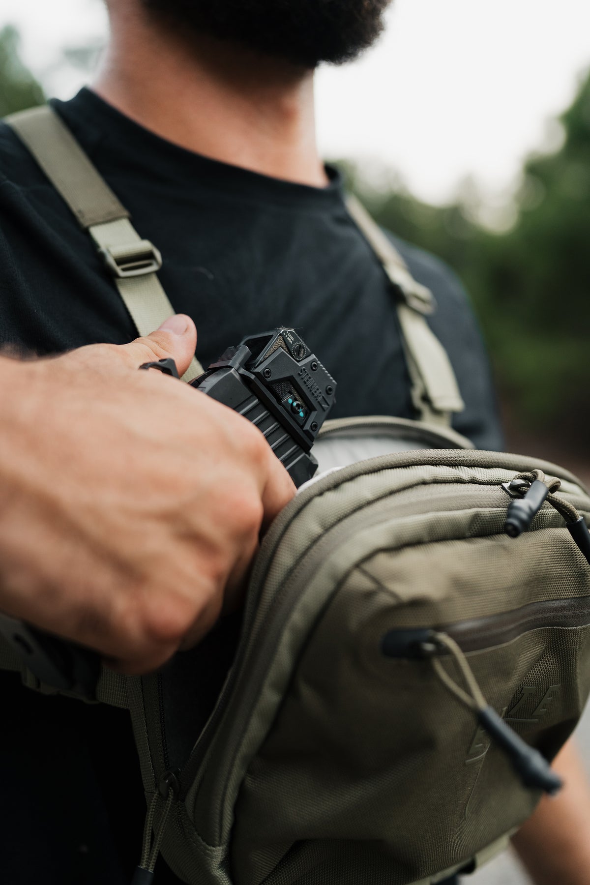 A close-up lifestyle shot of a person wearing an olive green tactical crossbody bag and black t-shirt, holding a black device against the bag's front pocket.