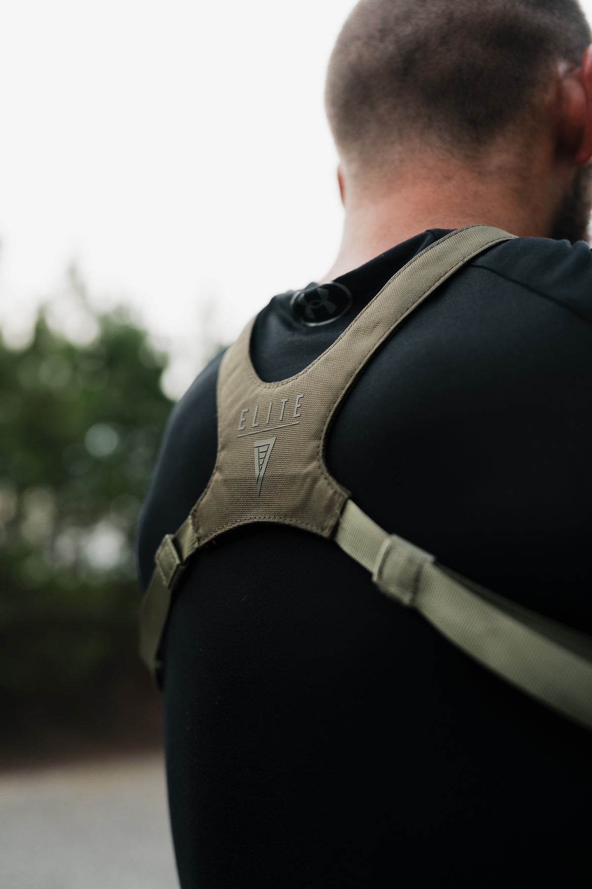 A close-up back view of a man wearing a tan tactical shoulder harness over a black shirt, with the brand name ELITE visible on the shoulder strap.