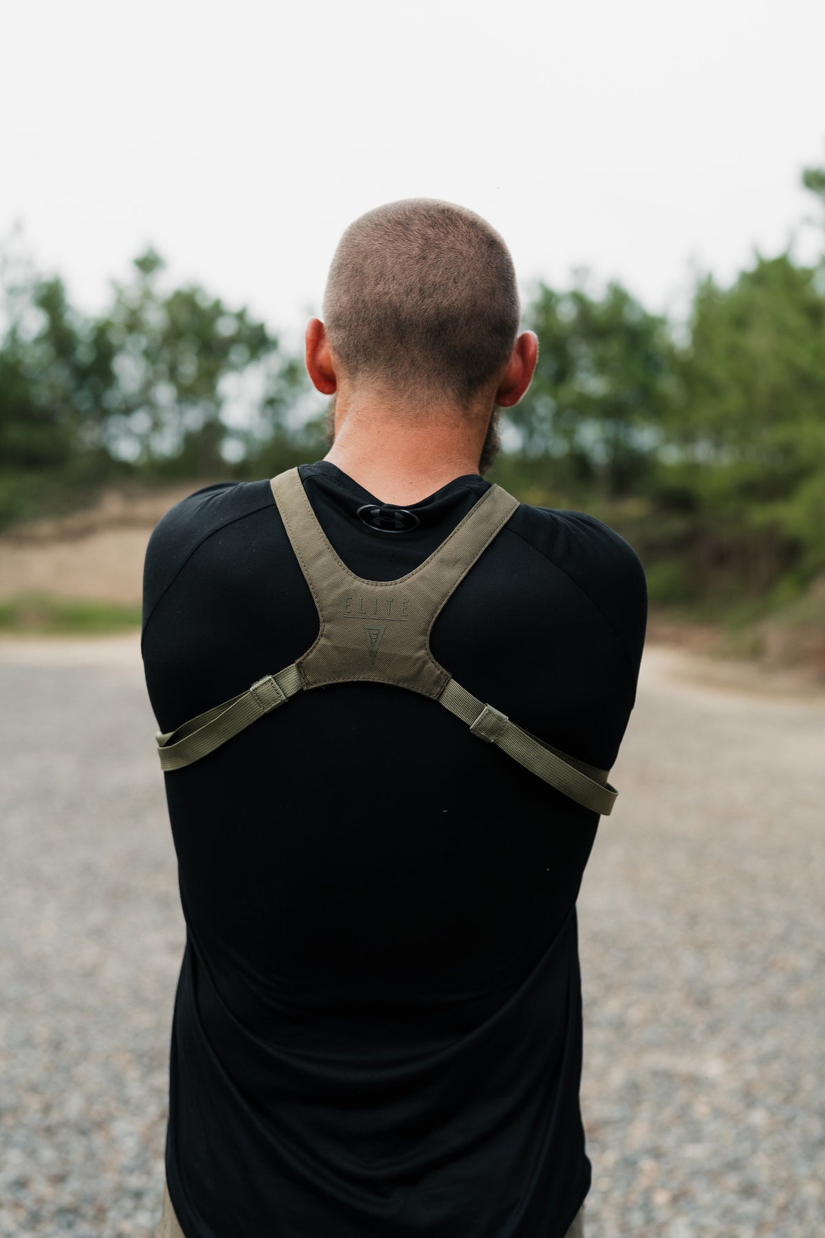 A man viewed from behind wearing a black athletic shirt and an olive green underarm support brace on his shoulders, standing outdoors on a gravel path with trees in the background.