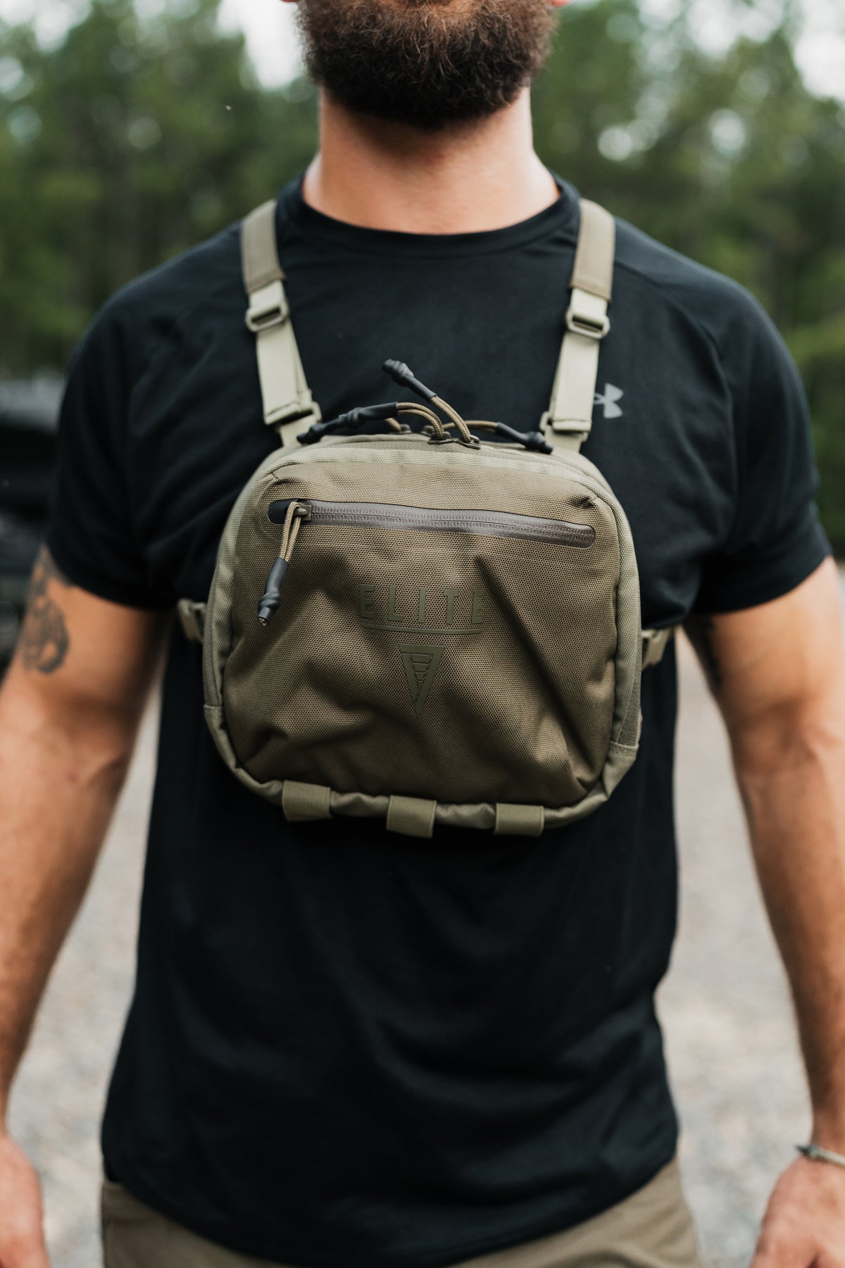 A man wearing a black t-shirt and khaki pants, shown from the chest down, wearing an olive green tactical chest bag with adjustable shoulder straps and a front zippered pocket.