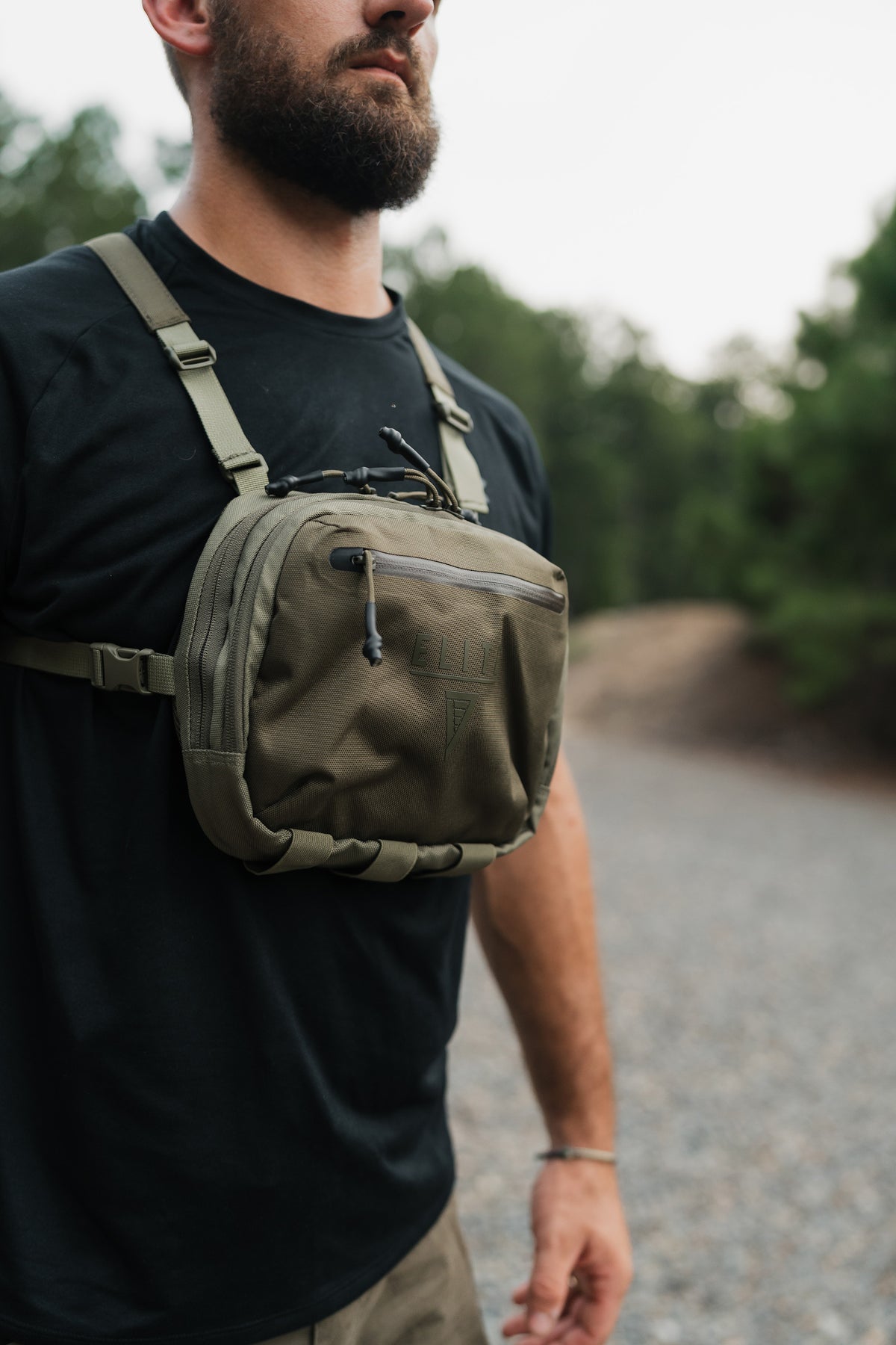 A man wearing a black t-shirt and khaki shorts carries a green tactical chest bag with a front zippered pocket and adjustable shoulder straps in an outdoor setting.