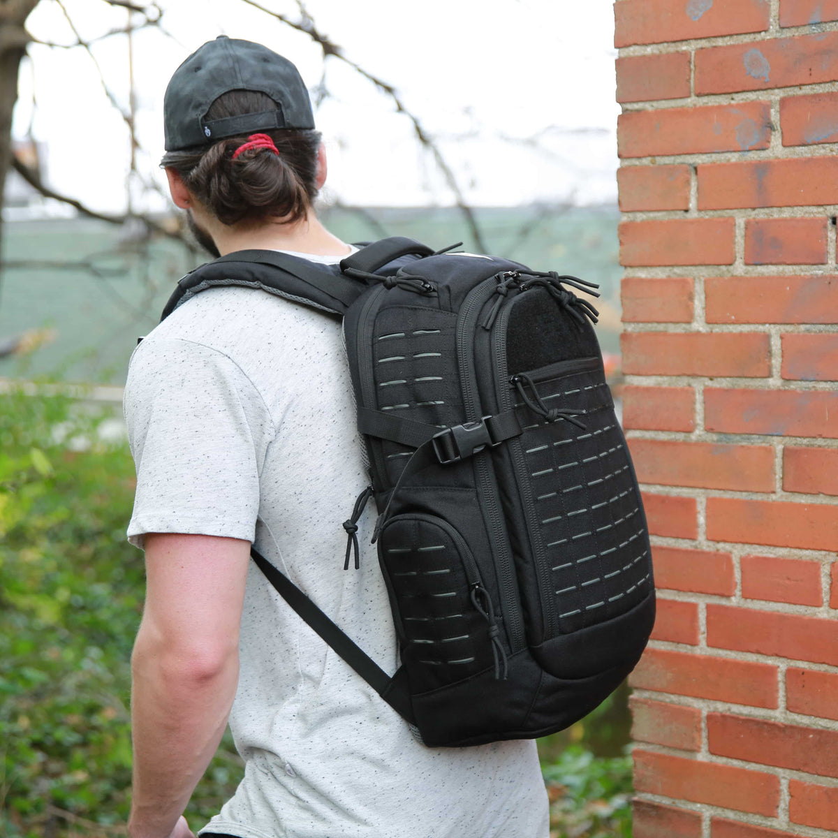 A man wearing a gray t-shirt and a dark cap, viewed from behind as he carries a black tactical backpack with a corrugated panel design, standing next to a red brick wall.