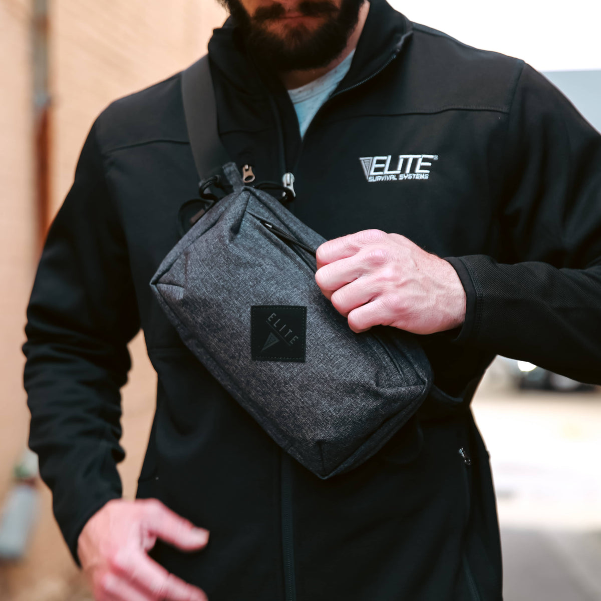 A man wearing a black Elite brand zip-up jacket and a dark gray heathered crossbody pouch bag with a black square logo patch, standing in an outdoor alleyway.