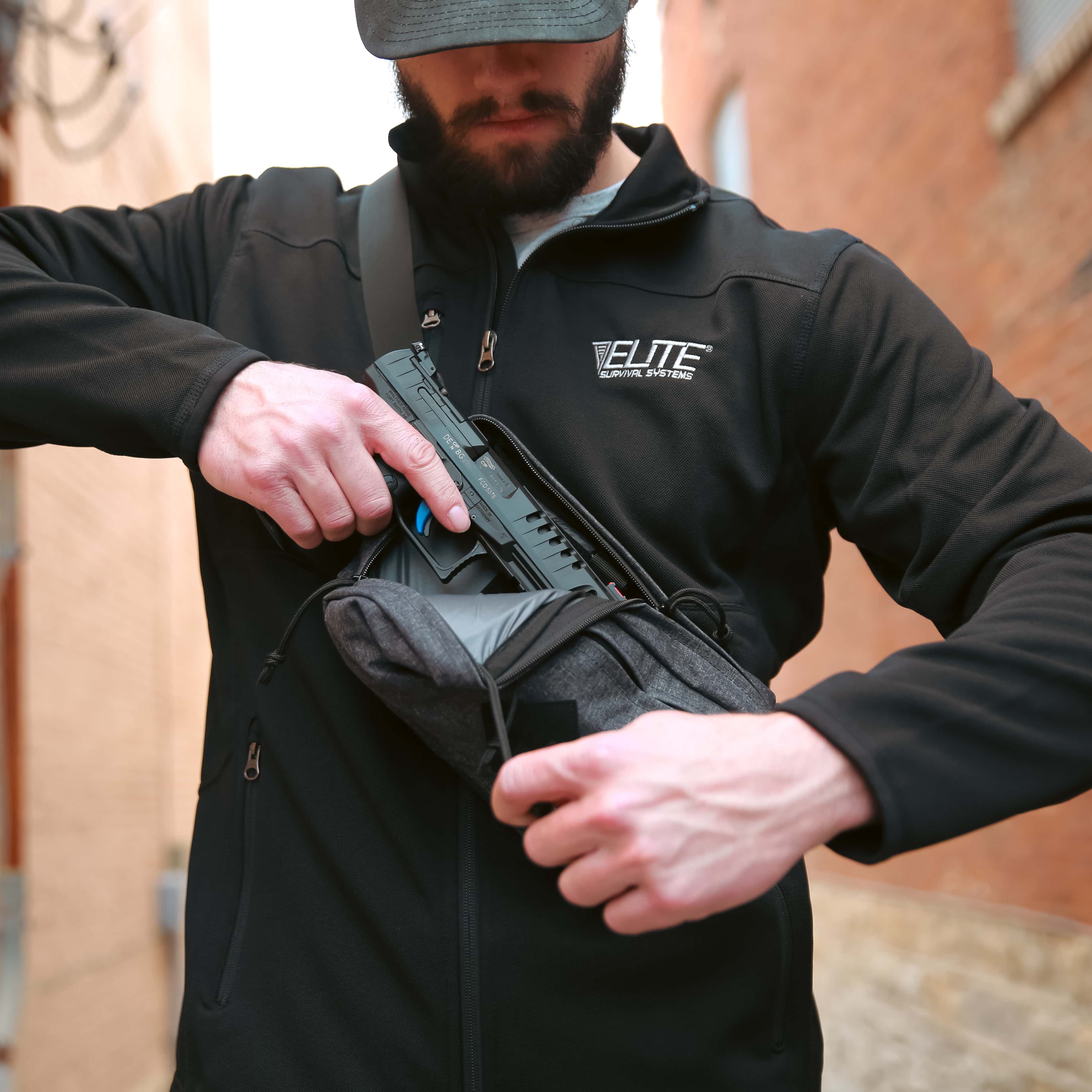 A man wearing a black zip-up fleece jacket with an 'ELITE' logo on the chest, shown from the waist up in an outdoor alley. He is demonstrating a black crossbody sling bag worn on his shoulder, opening the front pocket to show a handgun inside.