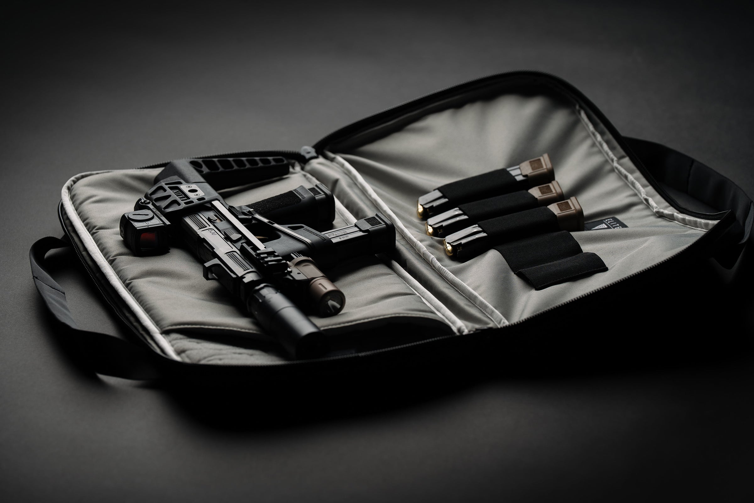 An open padded gun case on a dark studio background, holding a black rifle and several black cylindrical accessories with metallic tips.