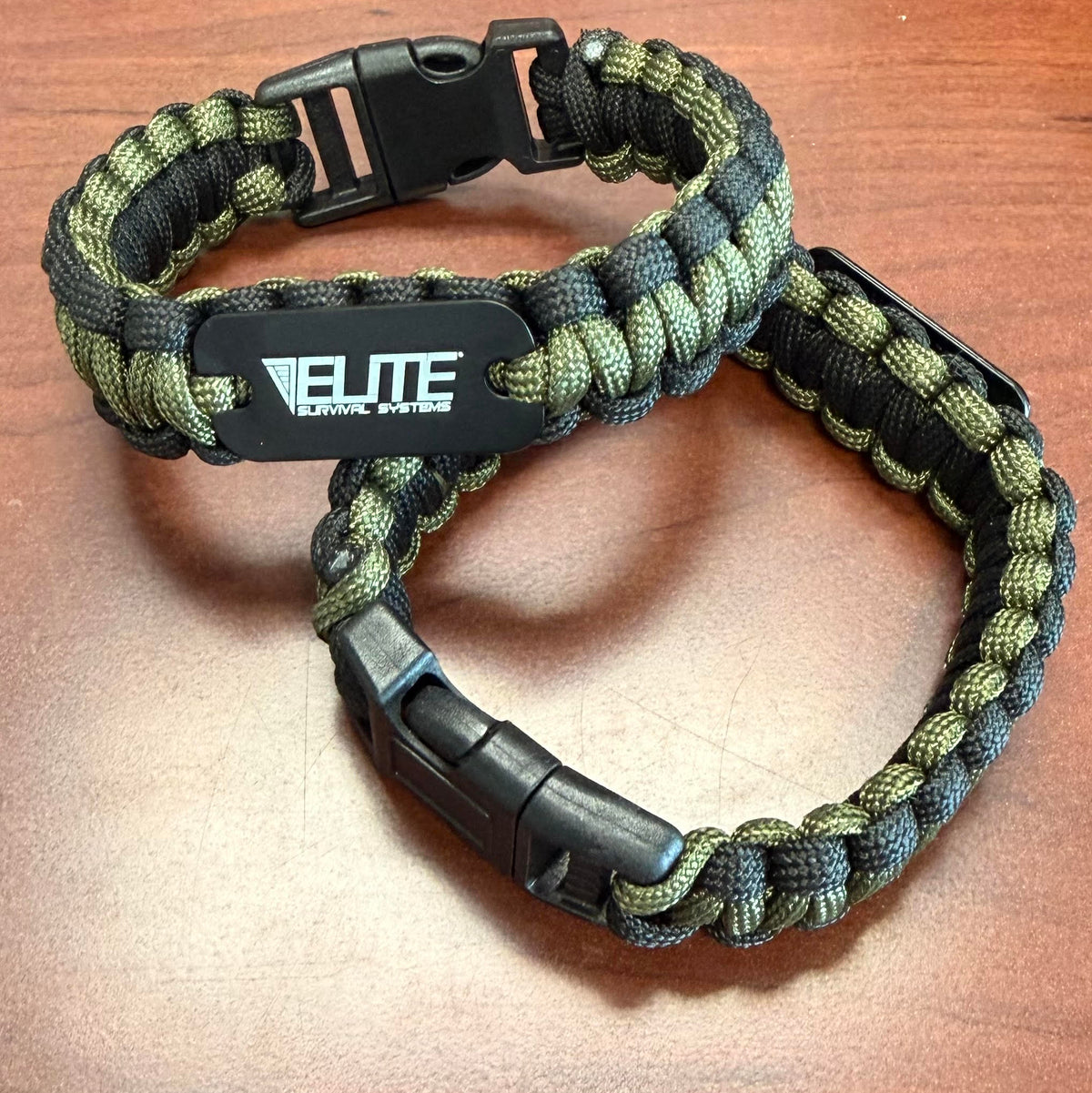 Two paracord survival bracelets in olive green and black, with one featuring a black plastic quick-release buckle and a central black plate printed with the brand name 'ELITE SURVIVAL SYSTEMS'.