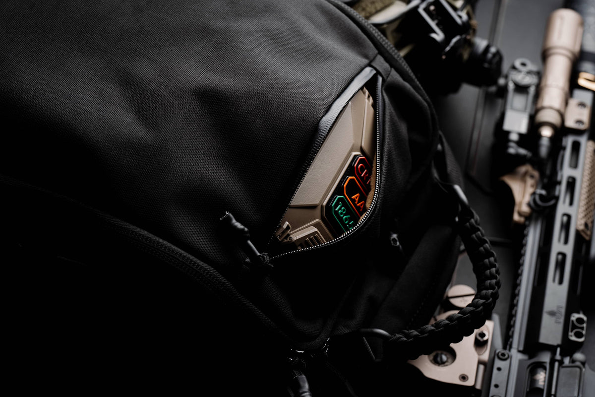 Zippered top pocket of the black Mission Backpack 40L open to show a handheld device, beside an AR-15 rifle